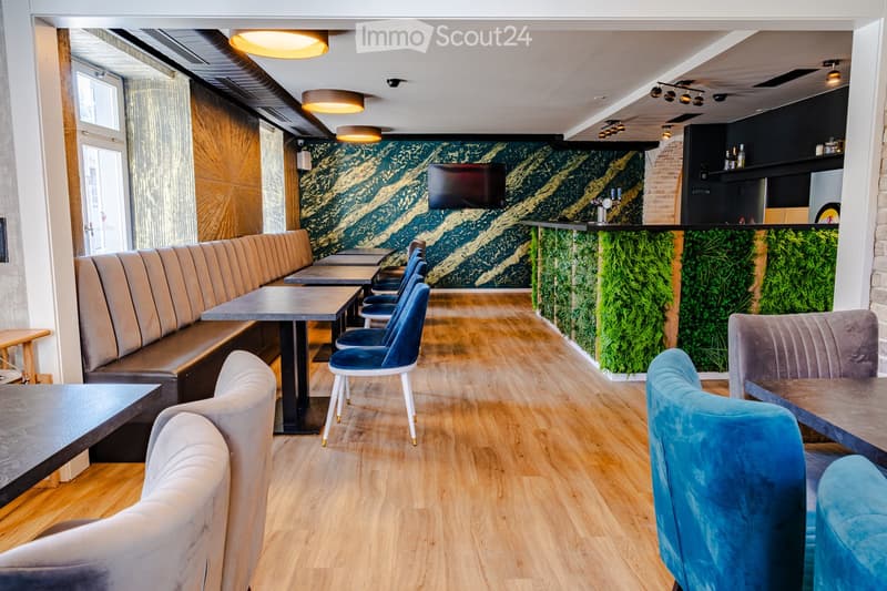 Modern restaurant interior with wooden flooring, upholstered seating, tables, and a television mounted on the wall.