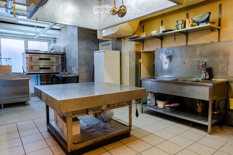 Commercial kitchen with stainless steel appliances, including a refrigerator, oven, sink, and a large central table. Shelves hold various kitchen items, and the space is well-lit with natural light from large windows.