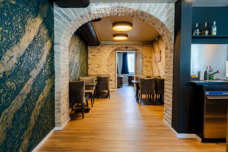 A stylish, well-lit room with exposed brick walls, wooden flooring, and modern furniture including tables and chairs. A small bar area is visible with a sink and bottles on the counter.
