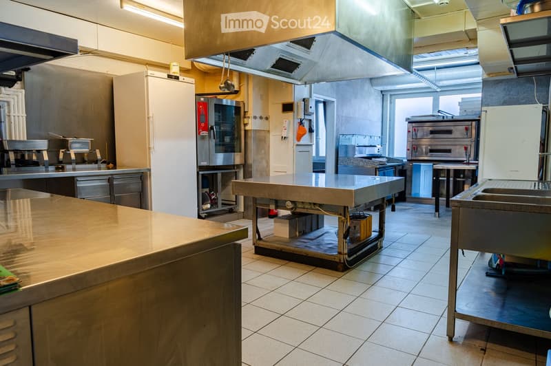 Commercial kitchen with stainless steel appliances, including a refrigerator, oven, sink, and counters. The space is well-lit with large windows and has tiled flooring.