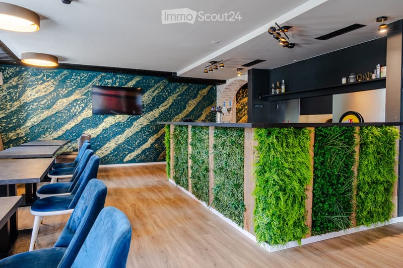 Modern interior with wooden flooring, blue upholstered chairs, tables, a bar area with bottles, and a green vertical garden wall.