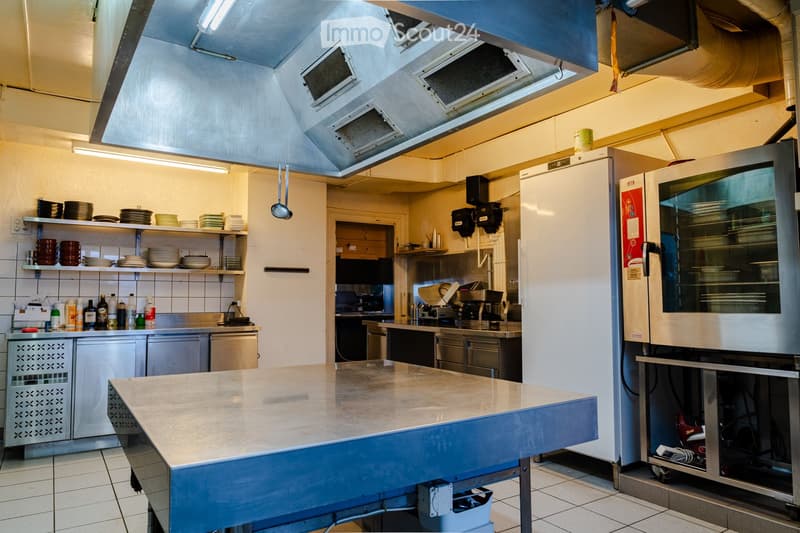 Commercial kitchen with stainless steel appliances, large central counter, shelves with plates and dishes, and various kitchen equipment.