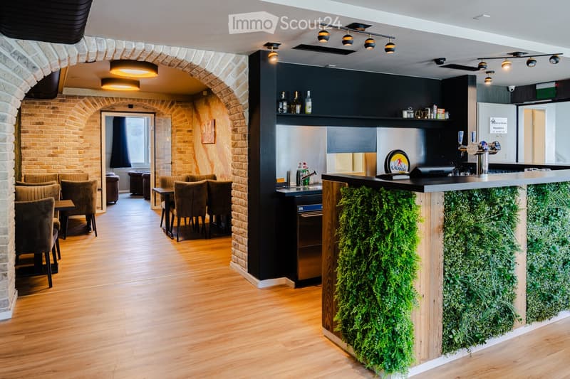 Modern interior with wooden flooring, brick walls, bar counter with green plants, seating area with sofas and tables, and a small kitchen area with a fridge and sink.