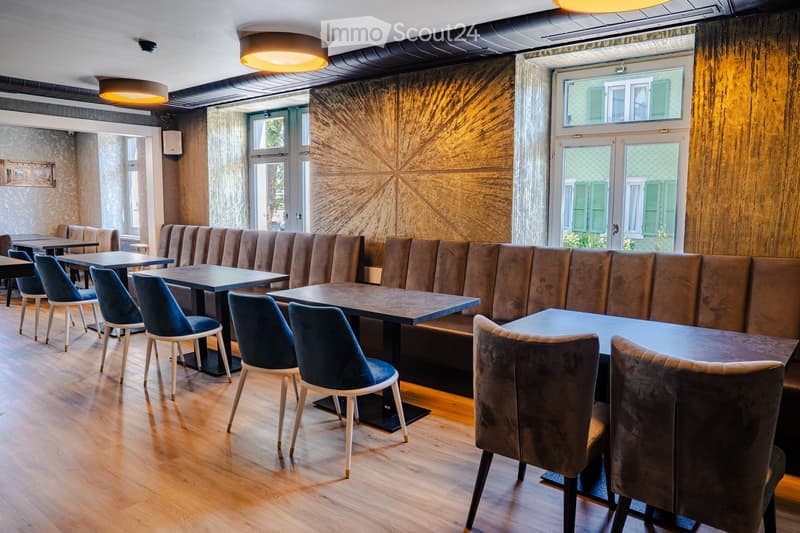 Modern restaurant interior with wooden floors, large windows, and contemporary furniture including tables, chairs, and a long bench seating area.