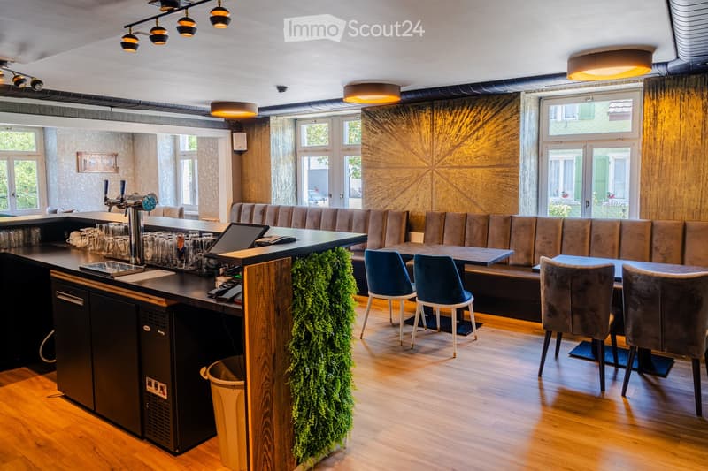 Modern bar area with a sleek black counter, bar stools, a long sofa, tables, and chairs. Features a built-in refrigerator, a faucet, and a laptop on the counter. The space is illuminated by warm, modern lighting and has large windows allowing natural ligh