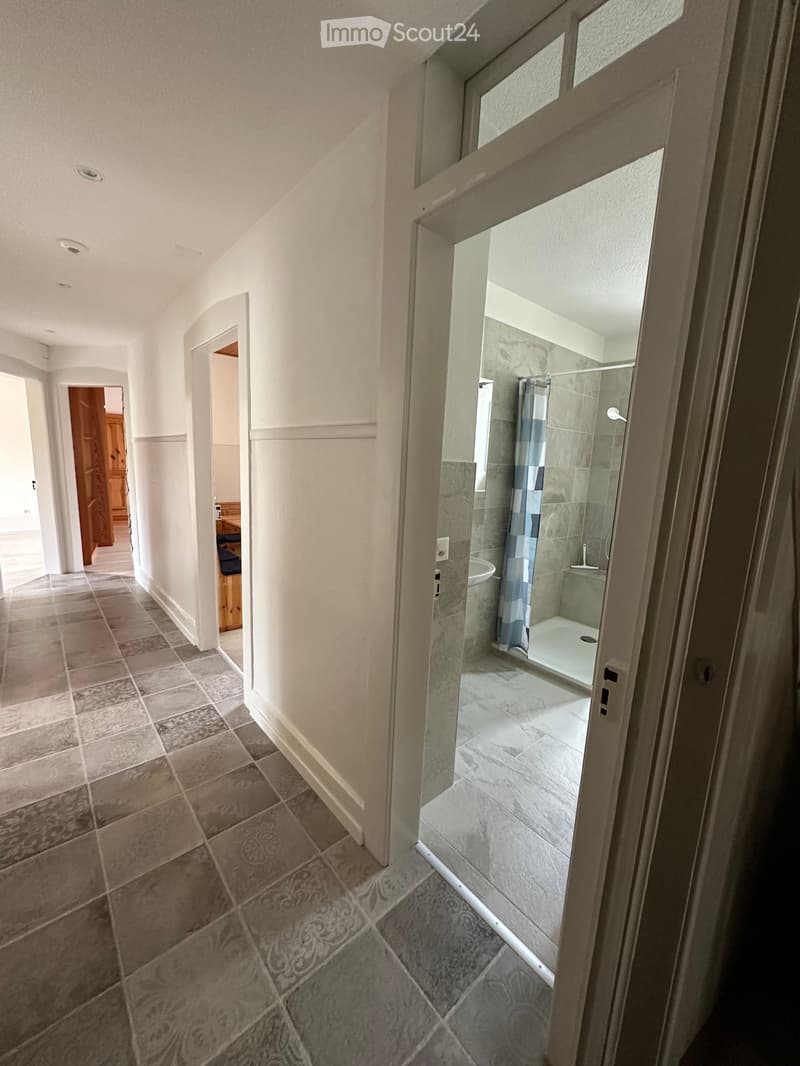 Corridor / Hallway. Empty corridor with tiled floor, white walls, and an open door leading to a bathroom.