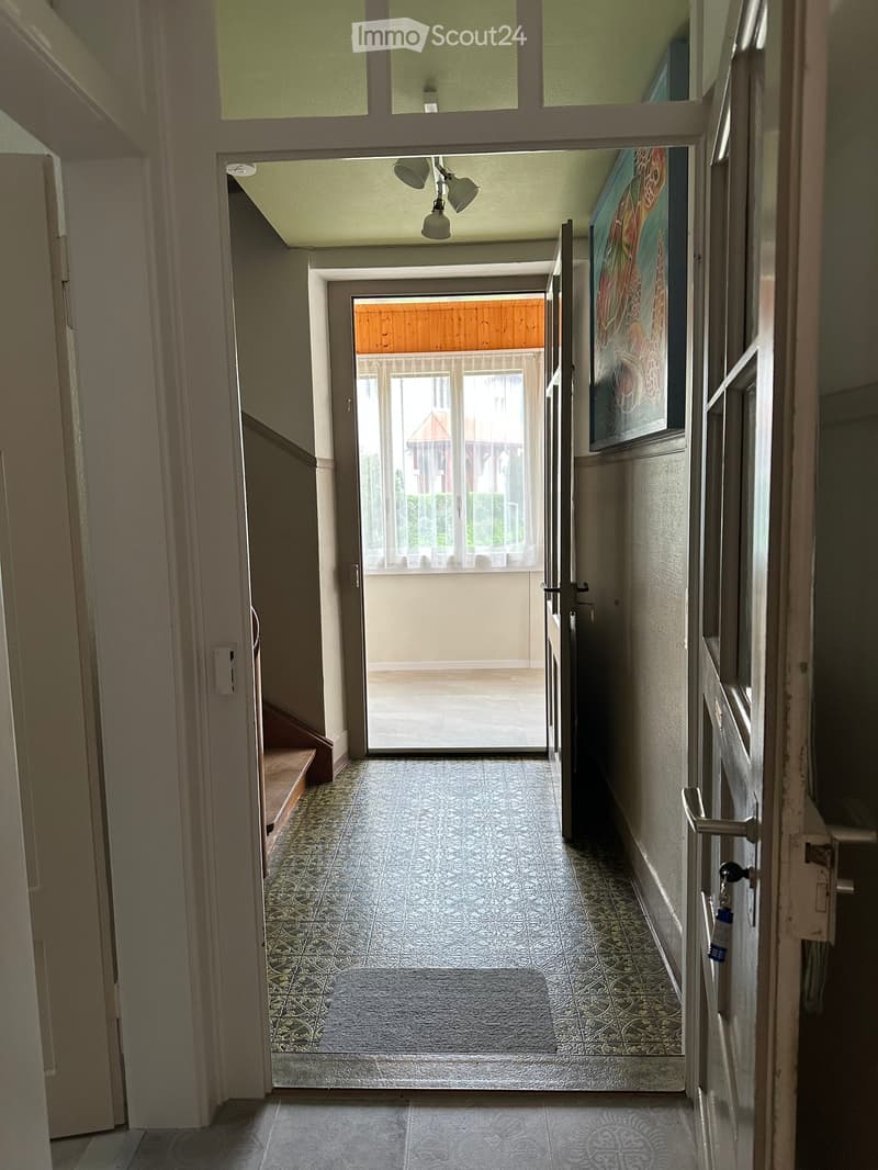 Corridor / Hallway. A hallway with tiled flooring, a rug, and a painting on the wall. The hallway leads to an open door with a view of a room with a window and a wooden staircase.