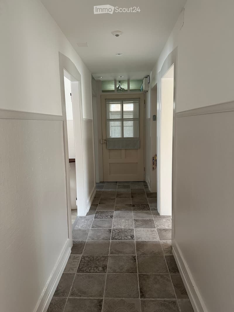 Corridor / Hallway. Empty hallway with tiled floor, white walls, and a door at the end.
