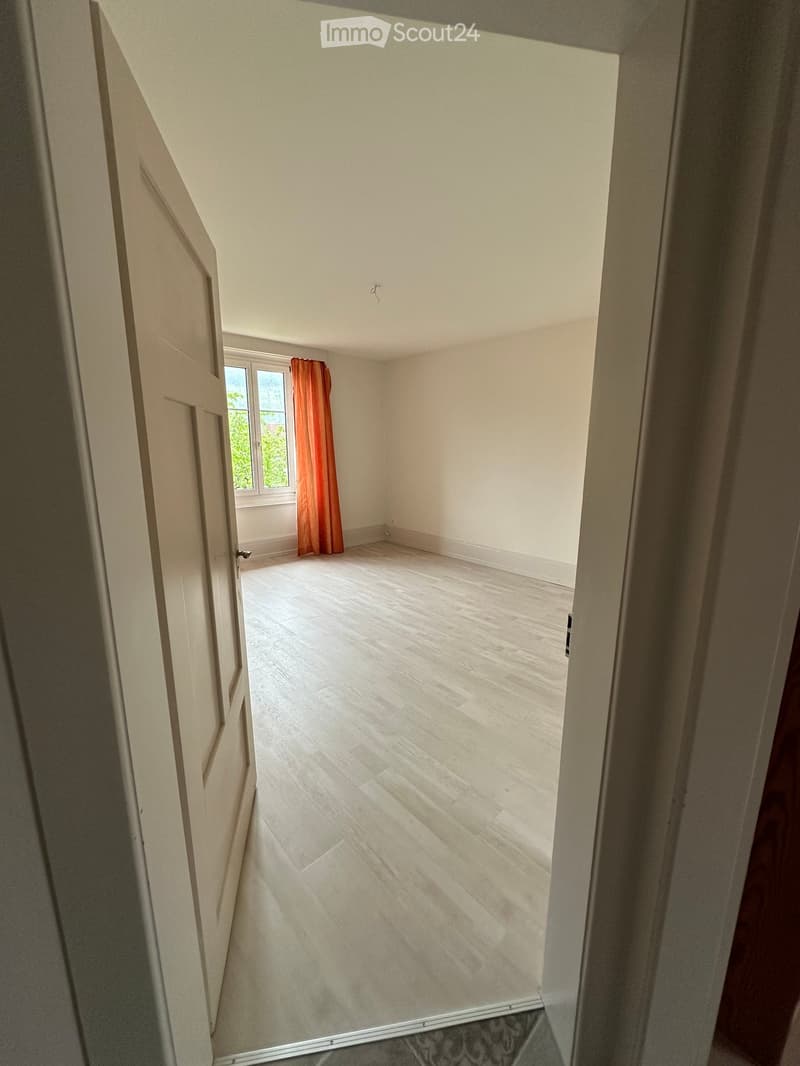 Unfurnished Room. An empty room with white walls, wooden flooring, and a window with orange curtains. The room has a door on the left side and a small object on the right side.