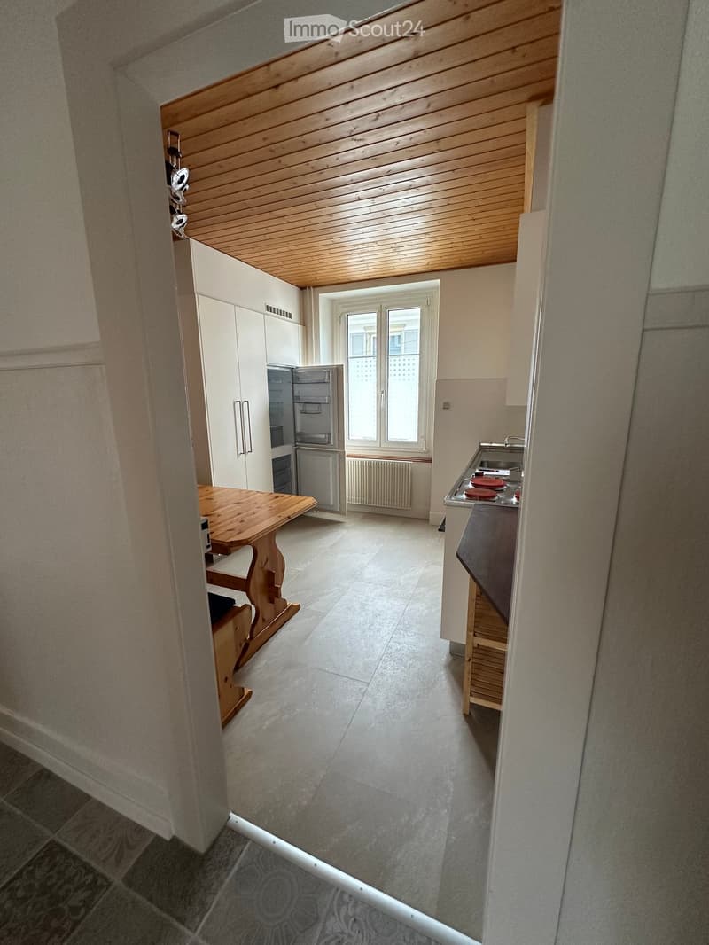 Kitchen. Open kitchen with wooden table, refrigerator, and stove. Tiled floor and white walls.