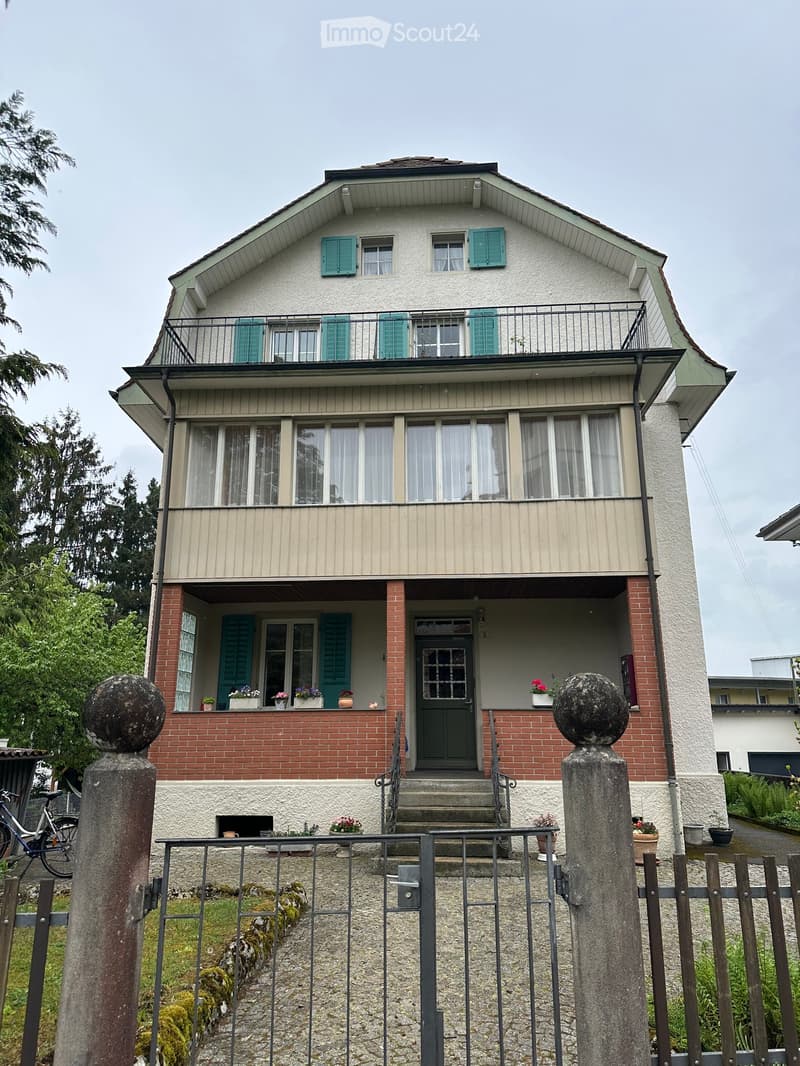 House. Two-story house with a brown roof, white and brick walls, green shutters, and a balcony. The entrance is flanked by two stone pillars with decorative tops. There are potted plants on the balcony and in front of the house. A bicycle is parked near t