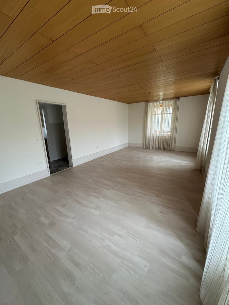 Unfurnished Room. An empty room with white walls, wooden ceiling, and wooden flooring. There are two doors, one of which leads to another room. There are two windows with white curtains. There are also two power outlets on the wall.