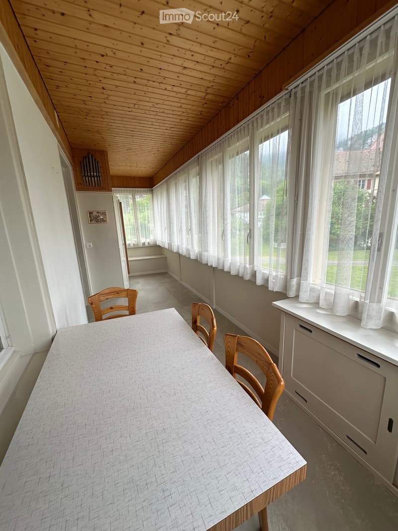 Furnished Room. A room with a wooden table and chairs, white walls, wooden ceiling, and large windows with white curtains. There is a cabinet with drawers under the window.