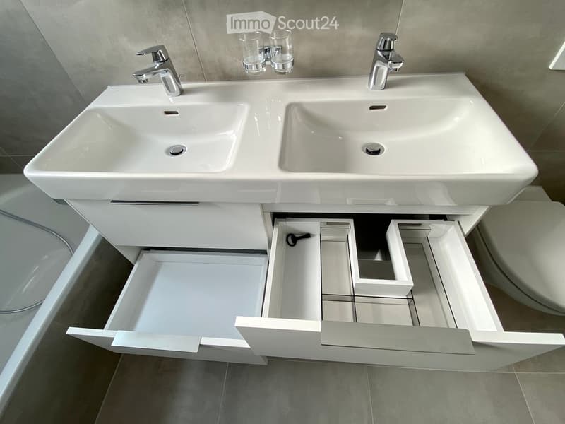 Modern bathroom with a double sink vanity, chrome faucets, and two clear cups placed on the countertop. The vanity has multiple open drawers and compartments.