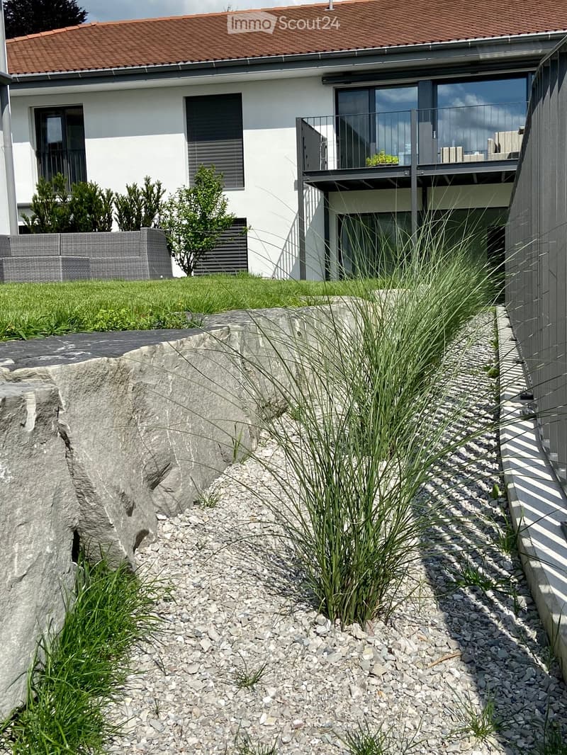 Modern house exterior with a raised ground floor, featuring a balcony, a well-maintained garden, and a stone retaining wall.