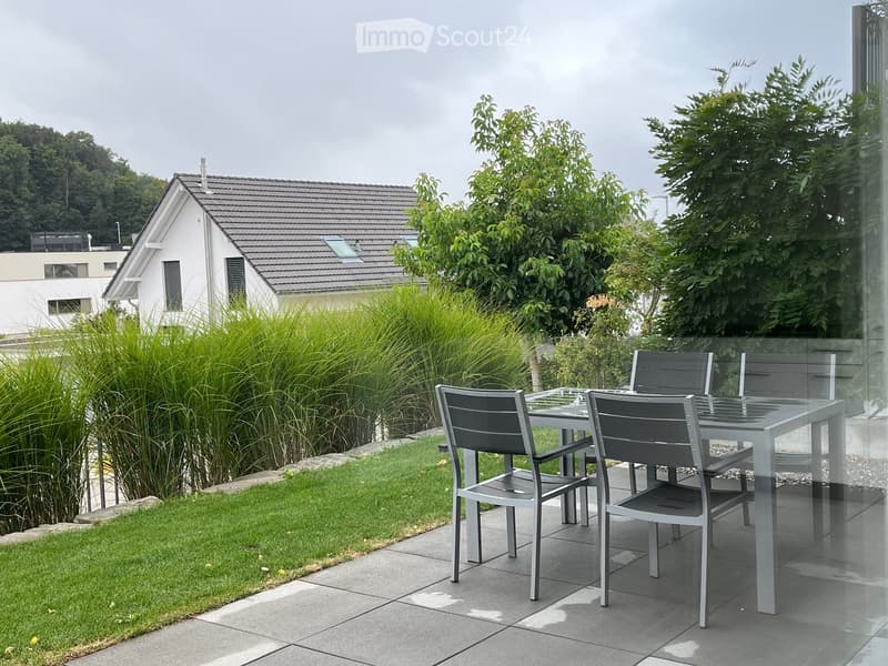 Outdoor patio with a glass table and four chairs, surrounded by a well-maintained lawn and tall plants, with a modern house in the background.