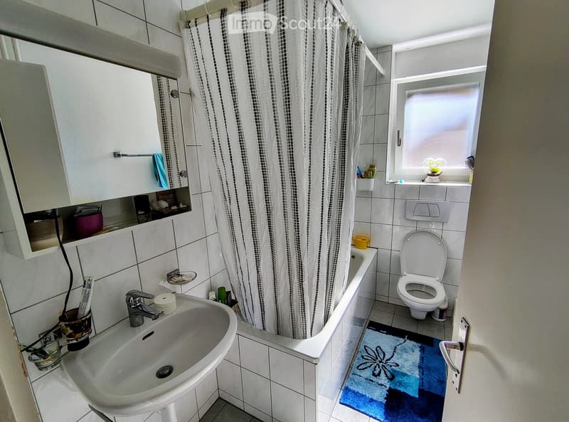 Bathroom with white tiles, a sink, a bathtub, a toilet, a mirror, a cabinet, a shower curtain, and a blue rug.