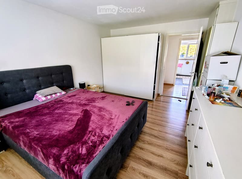 A modern bedroom with a large bed covered in a purple duvet, a nightstand with a lamp, a wardrobe, and a small table with a chair. The room has wooden flooring and white walls.