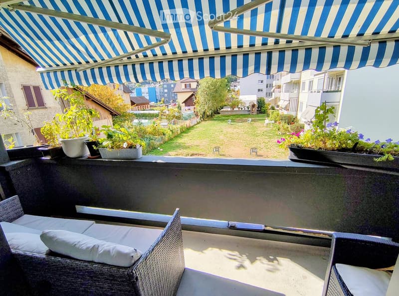 Balcony with a striped awning, sofa, table, chairs, and potted plants, overlooking a green garden area with additional seating and houses in the background.