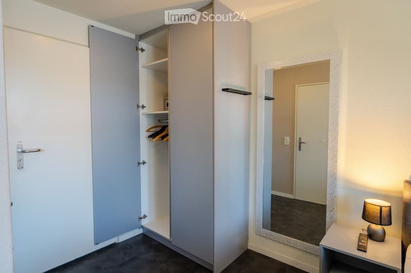 Modern, minimalist bedroom with a large mirrored wardrobe, a nightstand with a lamp, and a remote control on the nightstand.