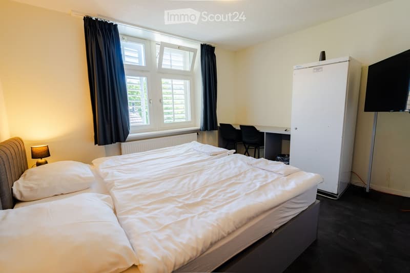 A well-lit bedroom with a double bed, a small refrigerator, a desk with chairs, a lamp, and a TV mounted on the wall. The room has large windows with dark curtains and a radiator below them.