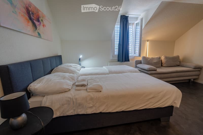 Cozy attic bedroom with a double bed, nightstand, sofa, and large window with curtains. The room features warm lighting and a modern aesthetic.