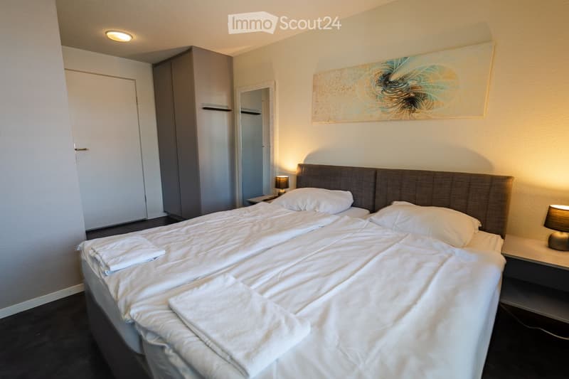 Modern bedroom with a large double bed, white linens, two nightstands with lamps, a wardrobe, and a decorative painting on the wall.