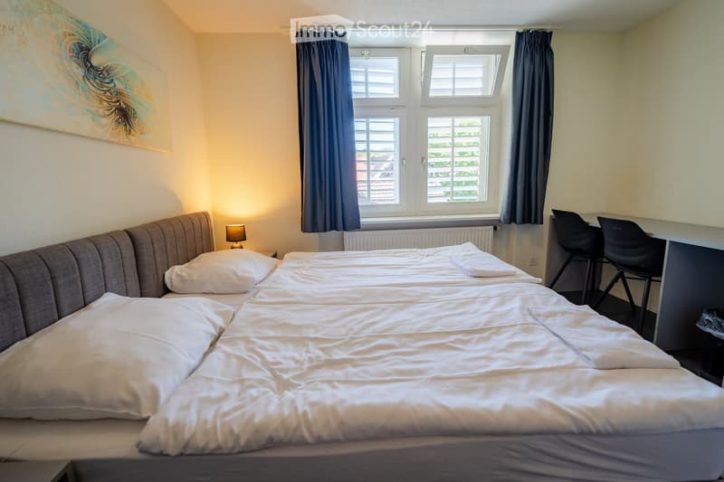 Cozy bedroom with two single beds, nightstand with lamp, desk with chairs, and large windows with curtains.