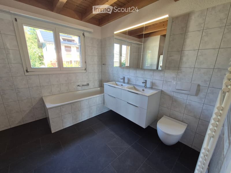 Modern bathroom with a bathtub, double sink, toilet, and large mirror. The room features tiled walls and a dark tiled floor, with a window providing natural light.