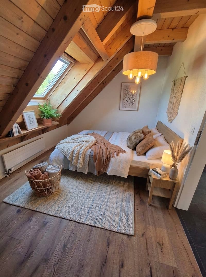 Cozy attic bedroom with wooden beams, skylight window, bed with pillows and blankets, nightstand with lamp, basket with towels, and decorative items.