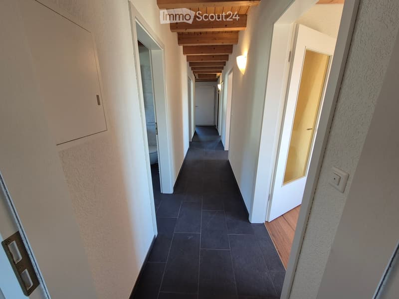 A long, narrow hallway with dark tiled flooring, white walls, and wooden ceiling beams. Several doors are visible along the sides, and the corridor is well-lit with wall-mounted lights.