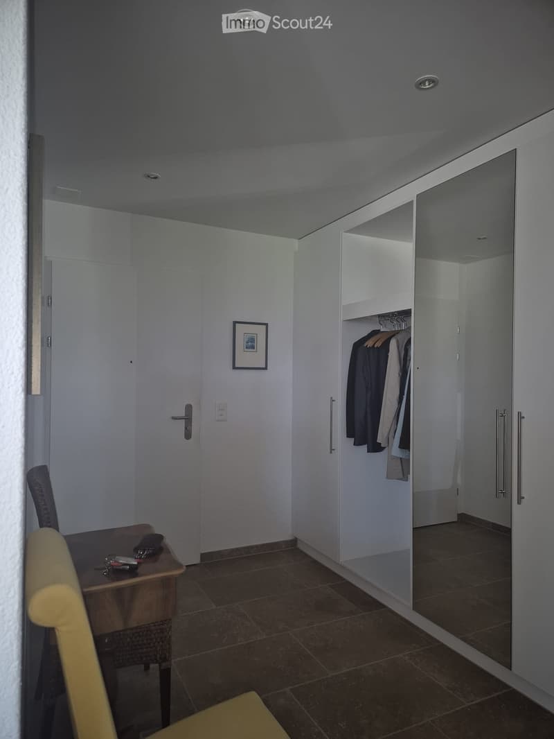 A small, well-lit room with a tiled floor, a wooden table with a chair, a wardrobe with hanging clothes, and a picture frame on the wall.