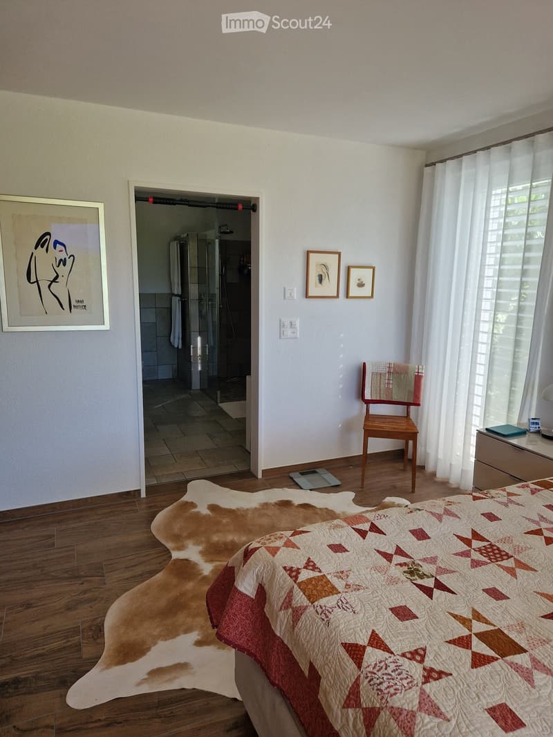 Bedroom with wooden flooring, a large bed with a patterned quilt, a cowhide rug, a wooden chair, framed artwork, and a small table with a digital scale and other items. An open doorway leads to a bathroom with a shower and towels.