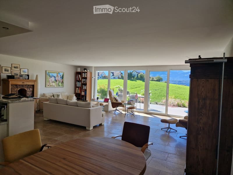 Bright living room with large windows offering a scenic view, featuring a cream-colored sofa, wooden coffee table, fireplace, bookshelf, and several chairs arranged around a central table.