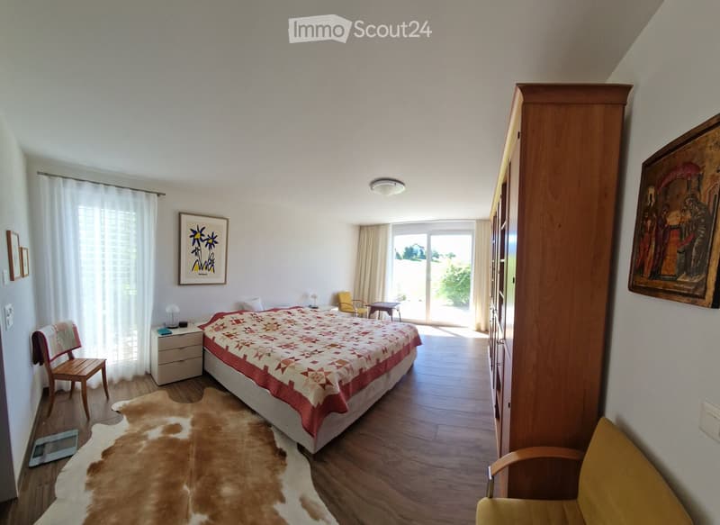 Bright bedroom with wooden flooring, large bed with patterned quilt, nightstand, chair, small table, wardrobe, and artwork on walls.
