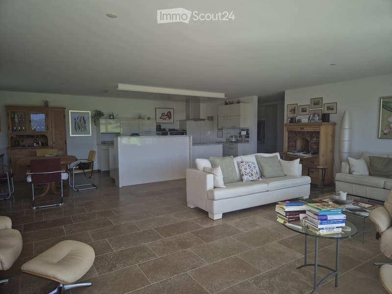 Modern living room with a white sofa, coffee table, armchairs, and a kitchen area in the background. The space features tiled flooring and contemporary decor.