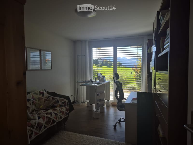 A cozy living room with a patterned sofa, a wooden table, a bookshelf, and a large window with blinds overlooking a scenic view.