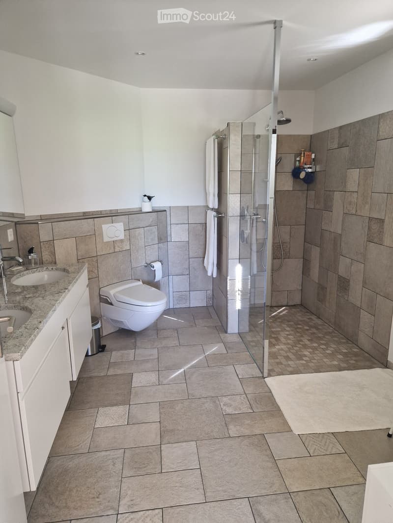 Modern bathroom with a glass-enclosed shower, toilet, sink, and tiled walls and floor. Includes a towel, cabinet, and a small trash bin.