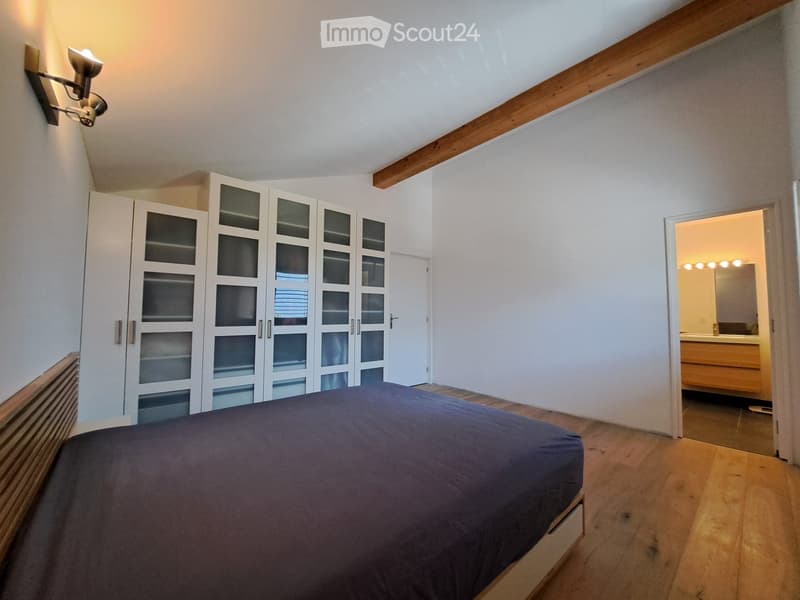 Bedroom with a dark blue bedspread, white wardrobe with glass doors, wooden floor, and an open door leading to a bathroom with a mirror and wooden vanity.