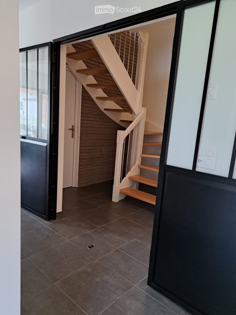 Modern hallway with a wooden staircase leading upwards, featuring a sleek black door frame and tiled floor.