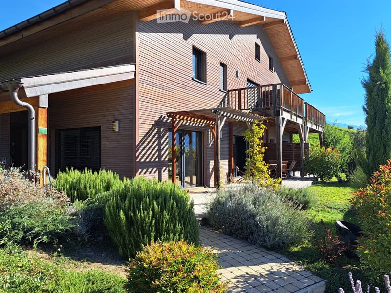 Two-story wooden house with a balcony, covered patio, and well-maintained garden with shrubs and a lawn.
