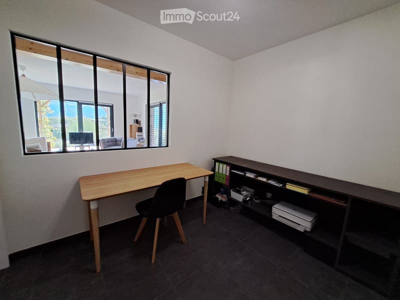 Modern office space with a wooden desk, black chair, shelving unit with books and office supplies, and a printer. Large windows provide natural light and a view of the outdoor area.