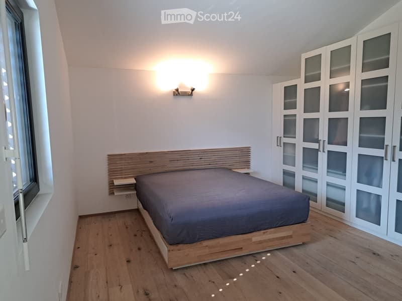 Modern bedroom with wooden floor, large window, bed with dark bedding, built-in wardrobe with glass doors, and wall-mounted lighting fixture.