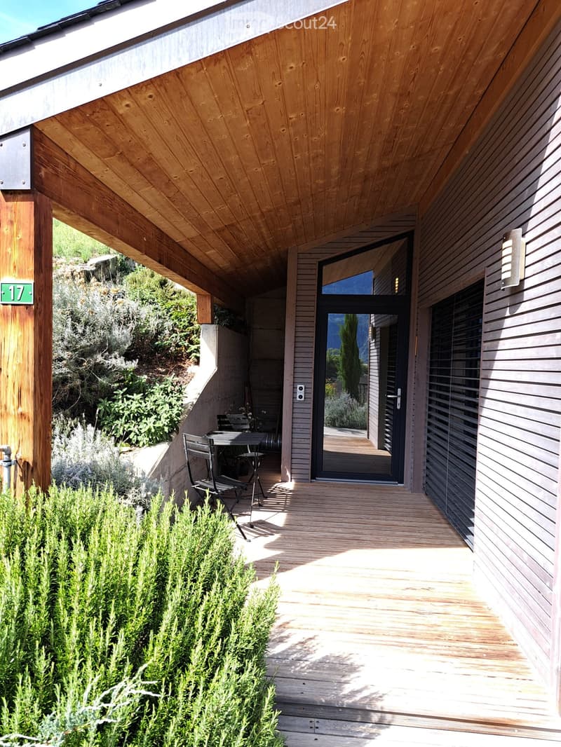 Wooden deck area with a table and chairs, surrounded by greenery and shrubs, with a modern house entrance visible in the background.