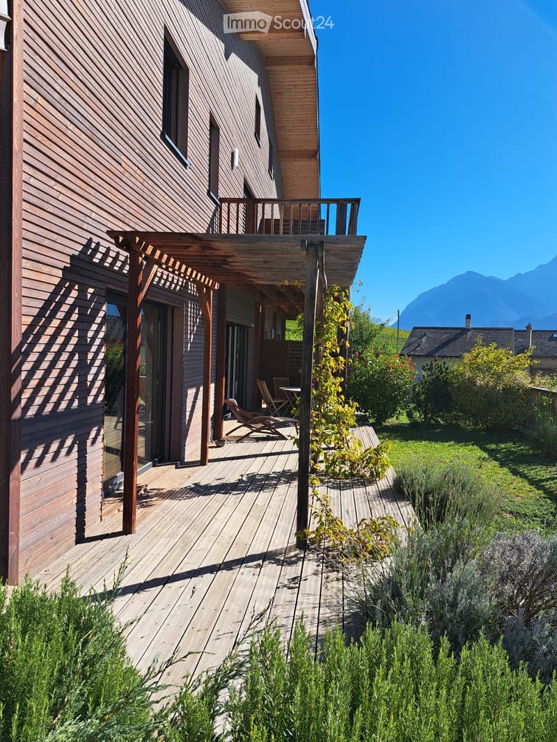 Wooden deck area with lounge chairs, small table, and surrounding greenery, with a modern wooden house in the background and mountain view.
