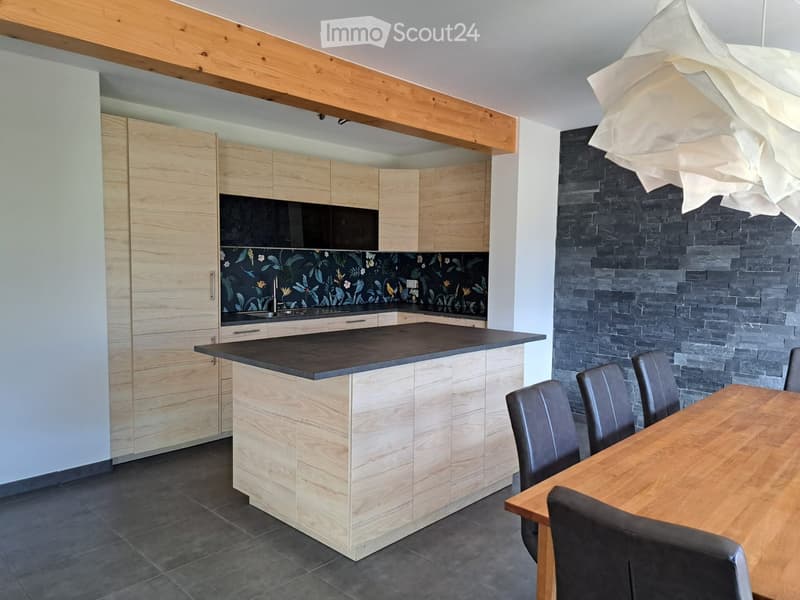 Modern kitchen with light wood cabinetry, dark countertops, and a central island. Features a dining area with a wooden table and black chairs, a decorative pendant light, and a floral backsplash.