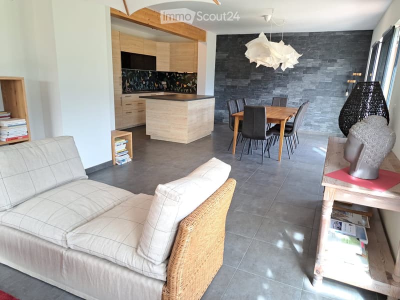 Open-plan living and dining area with a grey sofa, wicker chair, dining table with chairs, kitchen island, and wooden cabinets.