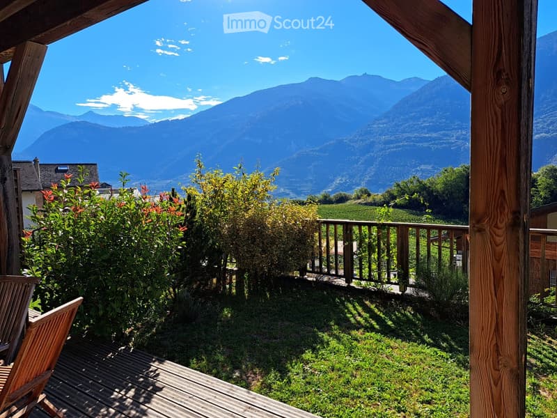 Balcony with wooden decking, wooden chairs, railing, and a view of mountains and greenery.