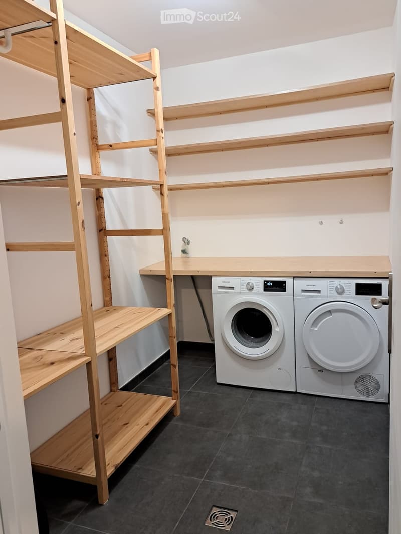 Modern bathroom with dark tiled floor, white walls, wooden shelves, and a washing machine and dryer unit.