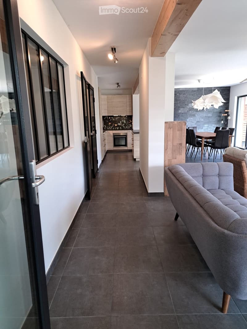 Modern hallway leading to a living area with a grey sofa, dining table with chairs, and a kitchen area with cabinets and an oven.